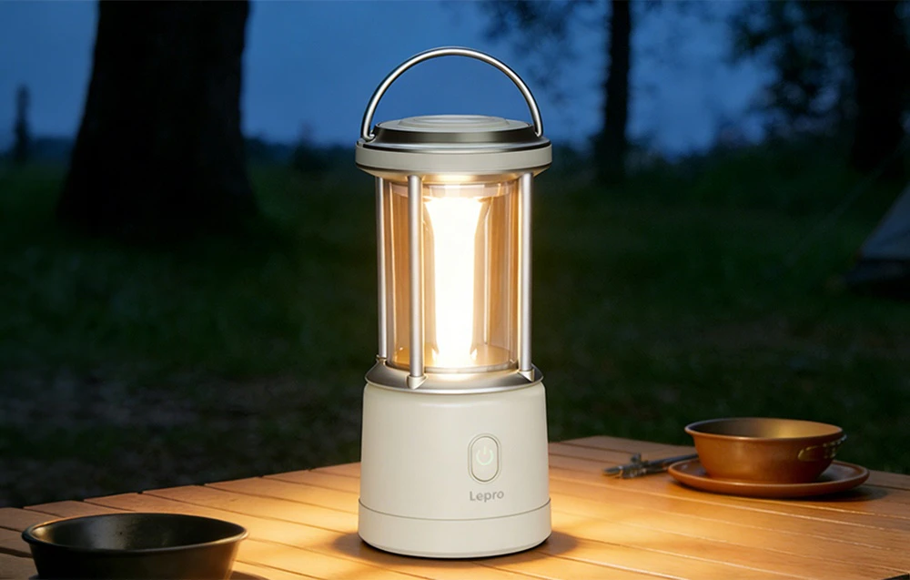 Lampa de camping Lepro aprinsă, așezată pe o masă de camping din lemn, pe fundalul unui peisaj de seară.