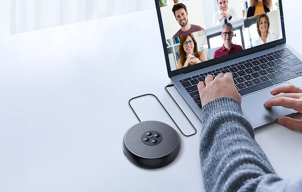 Un difuzor rotund pentru conferințe se află pe un birou alb și este conectat printr-un cablu USB la un laptop care afișează ferestrele participanților la videoconferință