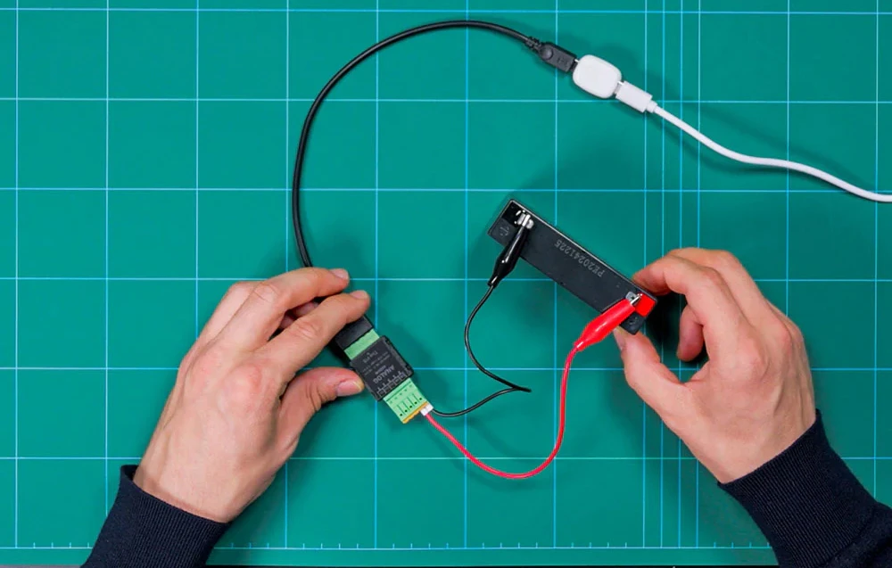 Două mâini care țin un modul negru cu un conector verde și o baterie cu numărul F232243224, conectate prin cabluri deasupra covorașului