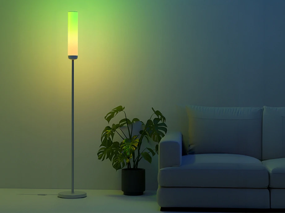 O lampă de podea modernă care emite lumină galben-verde, amplasată în sufragerie lângă o plantă monstera verde și o canapea de culoare deschisă.