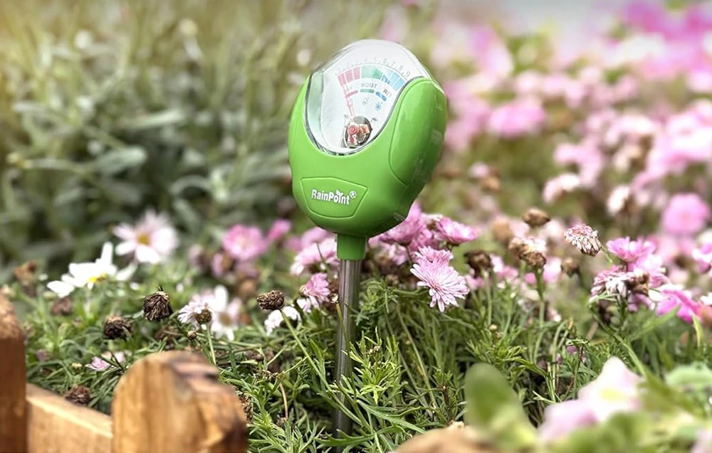 Umidimetru RainPoint verde pentru sol, amplasat printre florile roz înflorite din grădină
