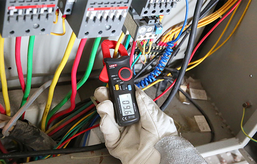 Clește ampermetric digital UNI-T în mâna unui electrician în timp ce măsoară curentul pe un fir într-un dulap de comandă.