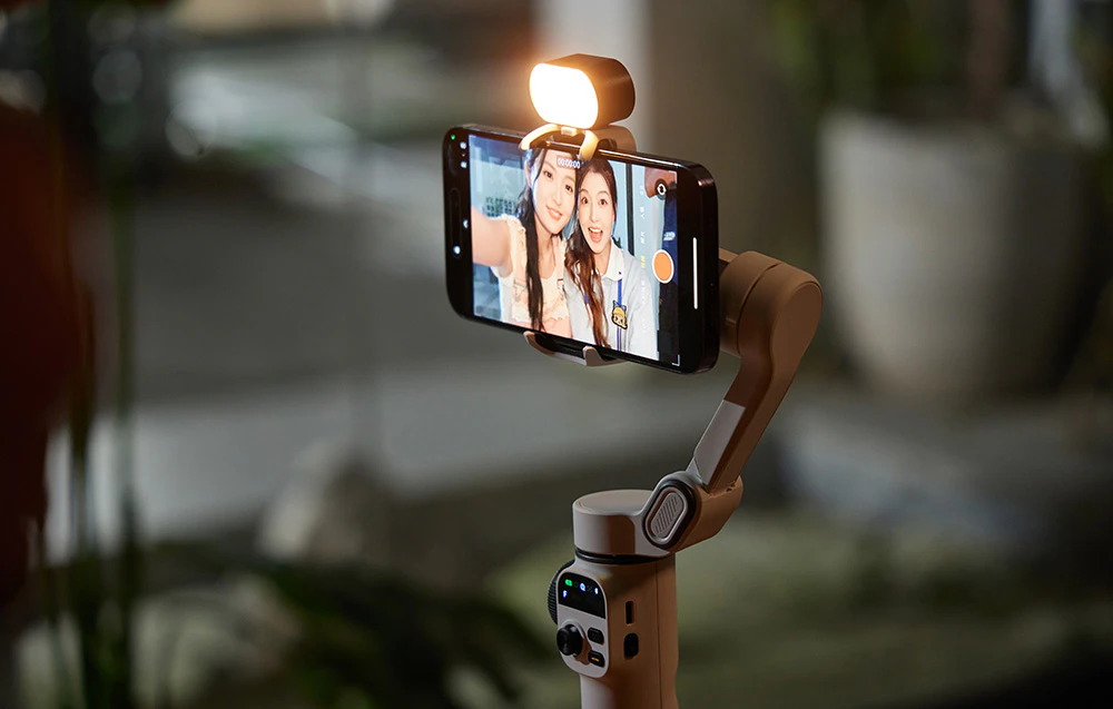 Un smartphone montat pe un gimbal bej cu lanterna aprinsă, afișând un selfie cu două femei.