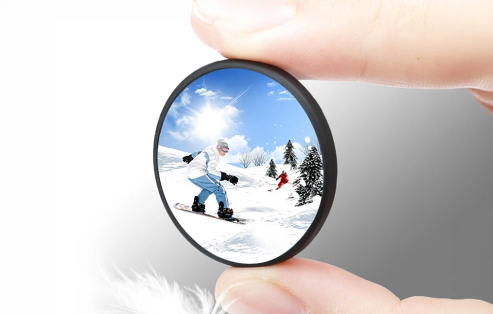 Degete ținând un capac rotund pentru lentile, în care se reflectă un snowboarder pe o pantă înzăpezită într-o zi însorită.