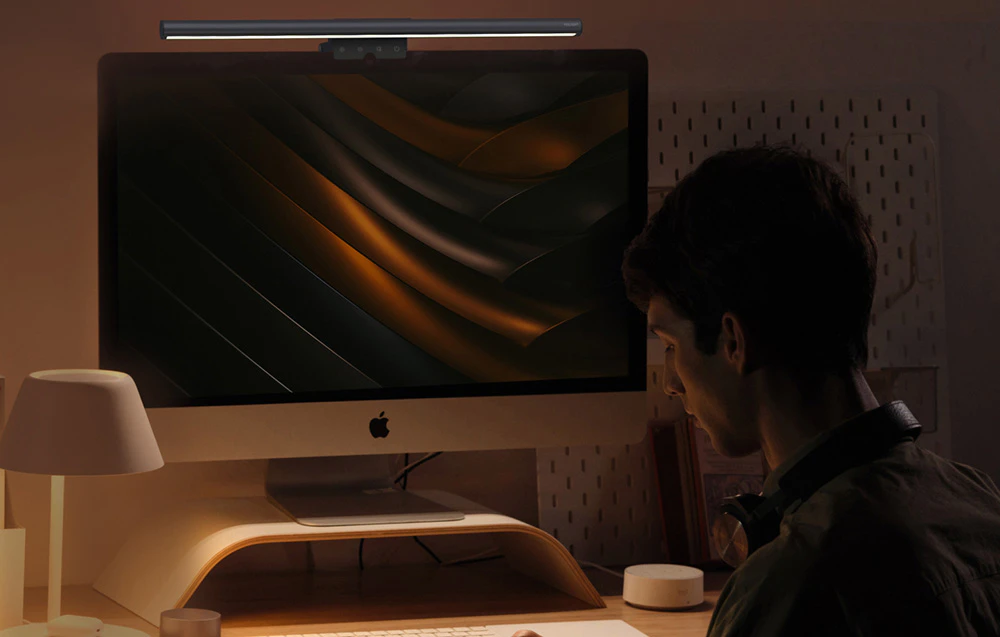 Un bărbat așezat în fața unui iMac cu o lampă montată pe partea superioară a ecranului, într-o cameră întunecată