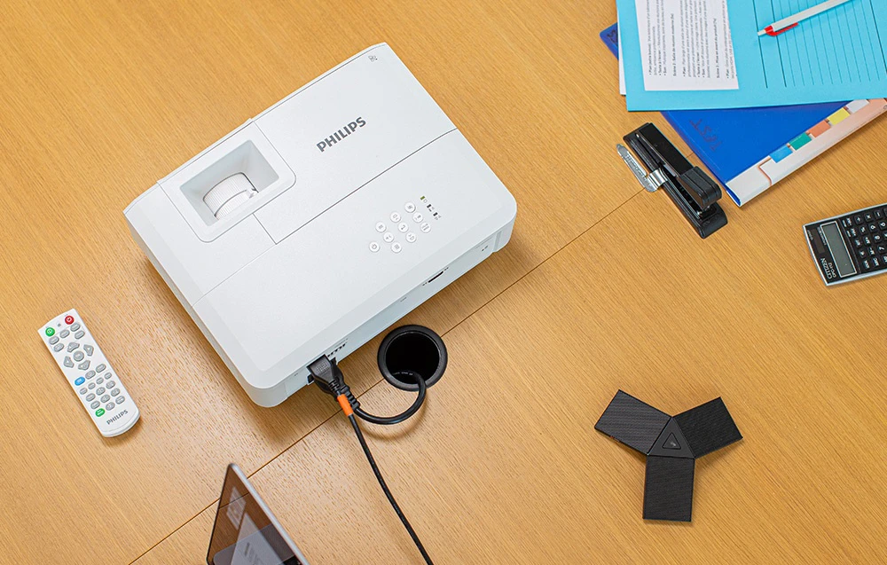 Vedere de sus a unui proiector PHILIPS conectat printr-un cablu la un port din blatul mesei, lângă o telecomandă și articole de birou