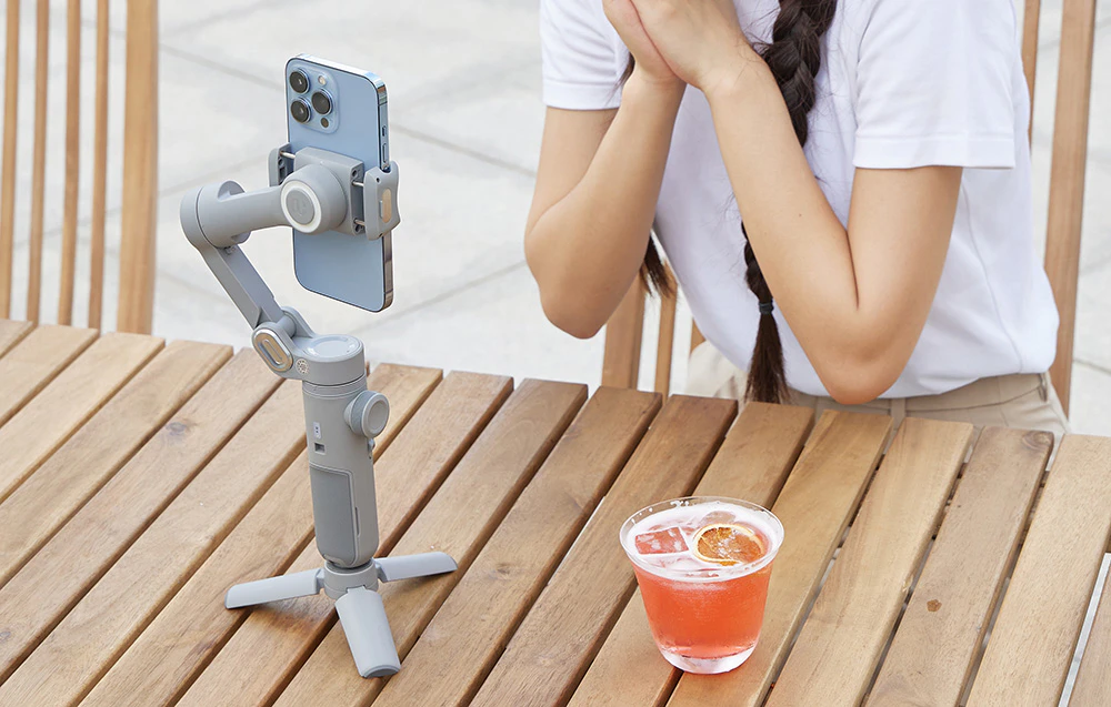 Un smartphone pe un gimbal montat pe un trepied înregistrează o imagine lângă o masă cu o băutură.