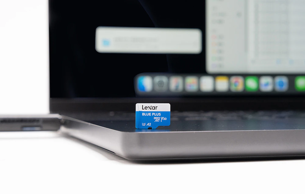 Prim-plan al cardului Lexar Blue Plus microSD așezat în poziție verticală pe carcasa unui laptop, cu un ecran estompat care afișează interfața sistemului vizibilă în fundal.