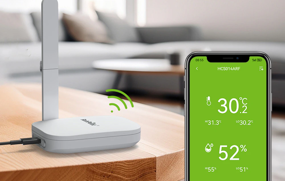 O stație de bază albă cu antenă se află pe o masă de lemn lângă un smartphone care afișează o temperatură de 30,2 °C și o umiditate de 52%