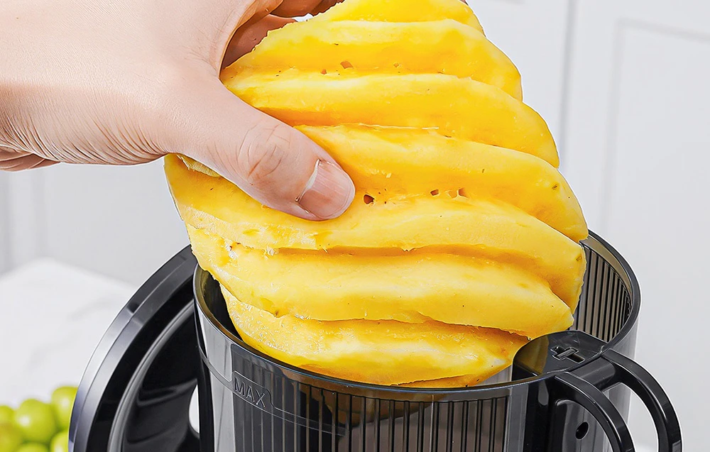 O mână introduce felii de ananas în recipientul storcătorului lent