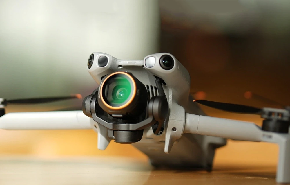 Dronă cu corp argintiu și cameră gimbal cu obiectiv verde și inel auriu pe masă de lemn