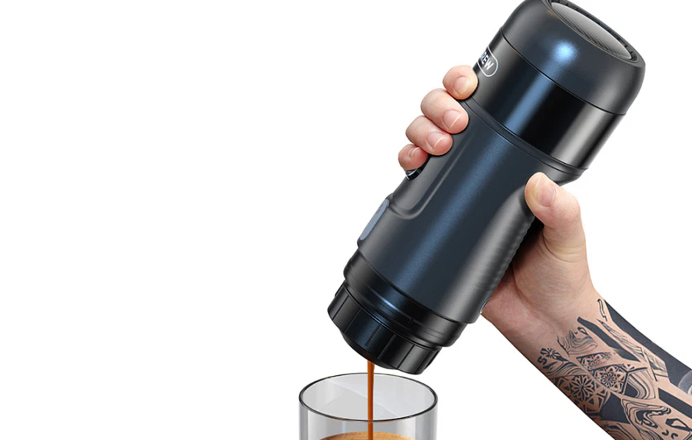 O persoană cu un tatuaj ține un aparat de cafea negru deasupra unui pahar în care se toarnă cafeaua.