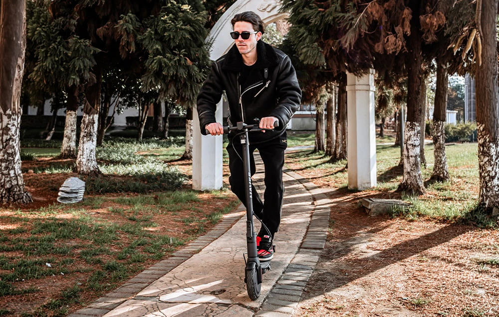 Un bărbat cu ochelari de soare conduce un scuter electric pe o alee din parc, printre copaci.