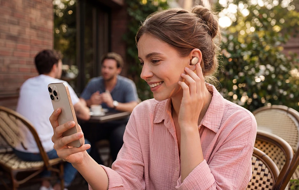 O femeie așezată în grădina unei cafenele folosește un smartphone și poartă o cască fără fir