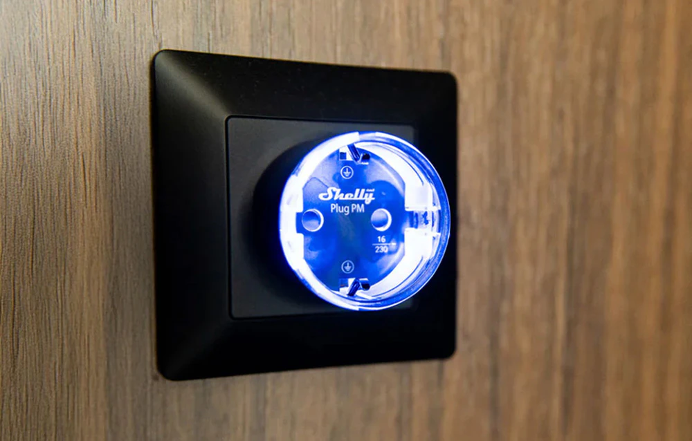 Priză inteligentă Shelly Plug PM cu iluminare LED albastră montată într-un cadru negru pe un perete din lemn.