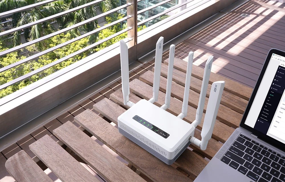 Un router alb cu șase antene se află pe o masă de terasă din lemn, lângă un laptop deschis