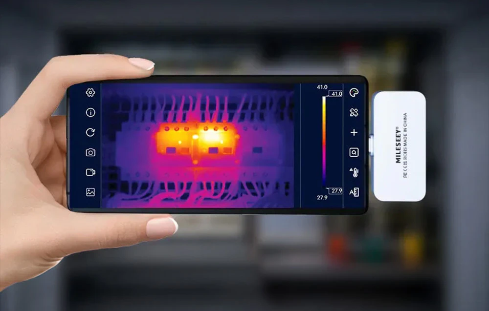 Un smartphone cu o cameră de termoviziune ținut în mână arată punctele cu temperatură ridicată dintr-un dulap de comandă.