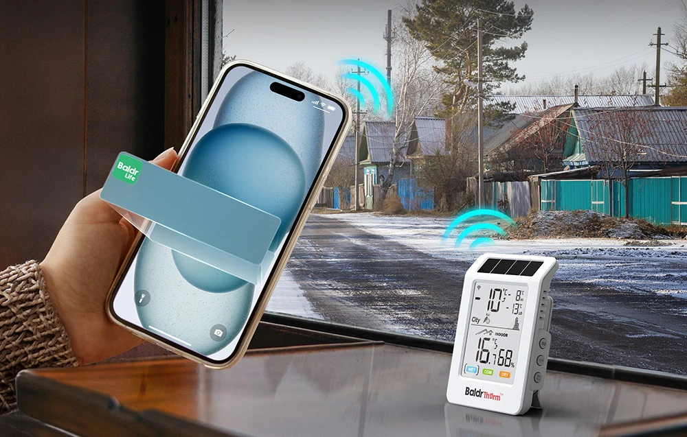 Un smartphone ținut în mână; lângă acesta, pe o masă, un termohigrometru BALDRTHERM