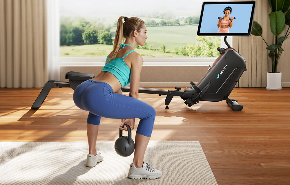 O femeie face genuflexiuni cu o greutate de tip kettlebell, iar în fundal se află un aparat de vâslit cu un ecran de antrenament.