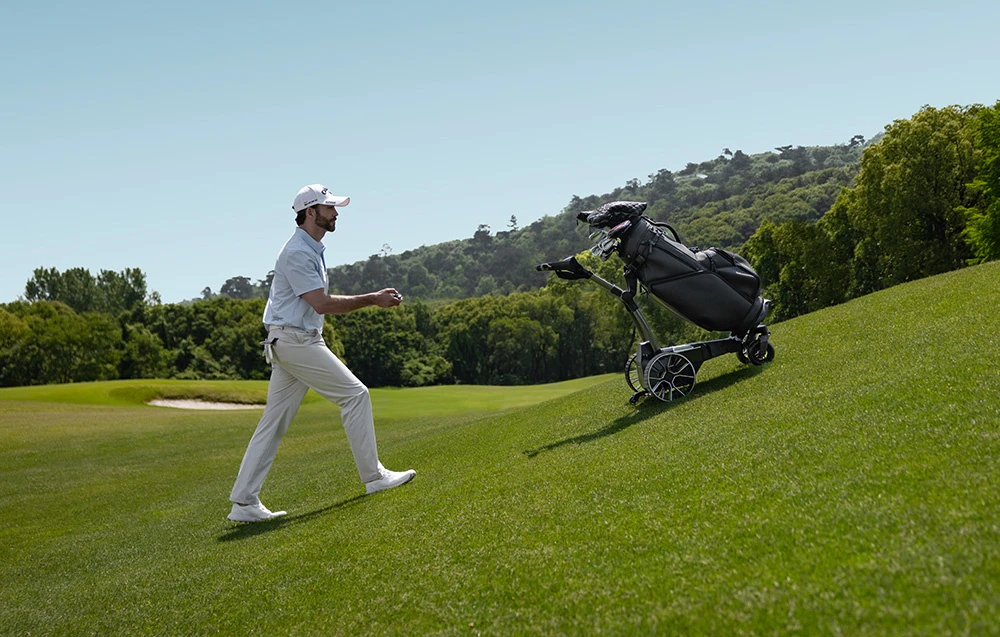 Un jucător de golf urcă dealul în spatele unei mașinuțe de golf electrice care transportă o geantă cu crose
