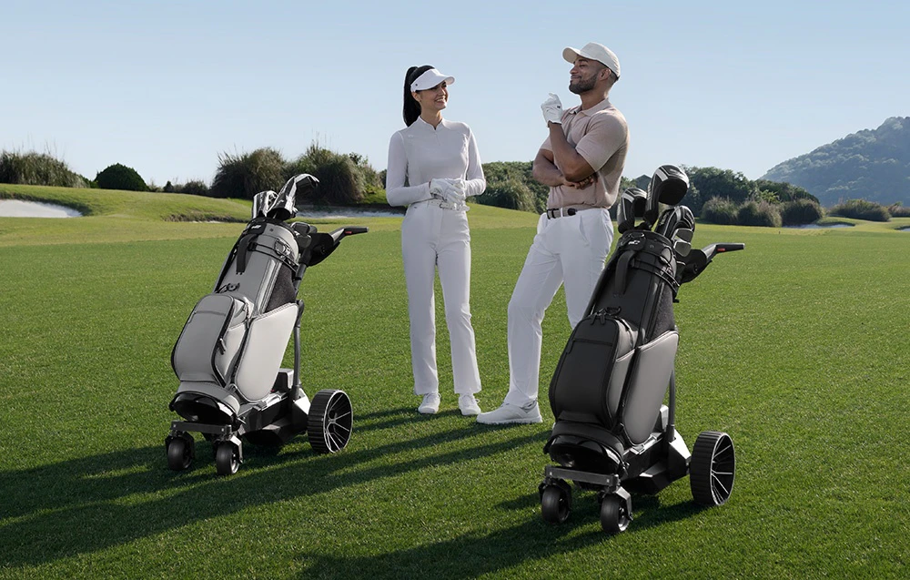 Doi jucători de golf discută pe terenul de golf, cu două cărucioare electrice cu saci de golf lângă ei