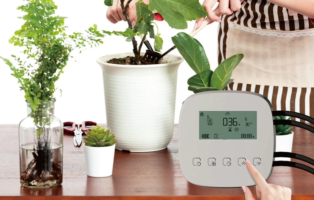 Mâini îngrijind o plantă în ghiveci, lângă un controler de irigare cu afișaj LCD și tuburi conectate