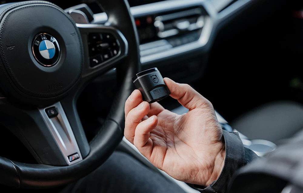 O mână ținând adaptorul OBD2 Carly în interiorul unei mașini BMW, cu volanul vizibil în fundal