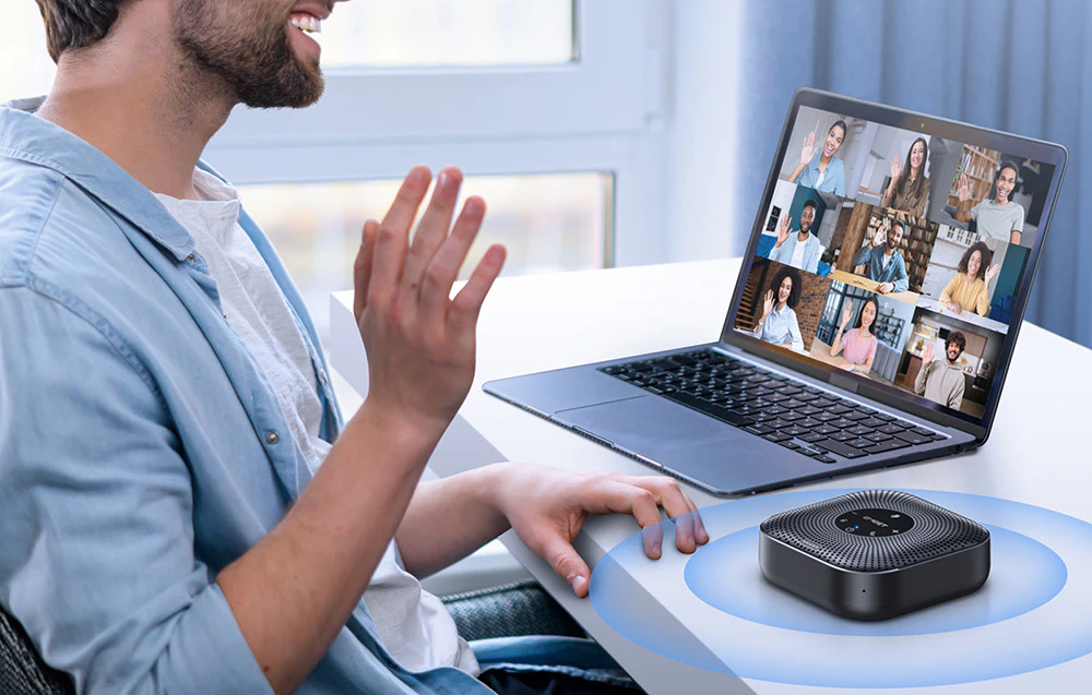 Un bărbat salută cu mâna participanții la videoconferință vizibili pe ecranul unui laptop, lângă care se află pe birou un difuzor negru, pătrat, pe care sunt marcate unde sonore