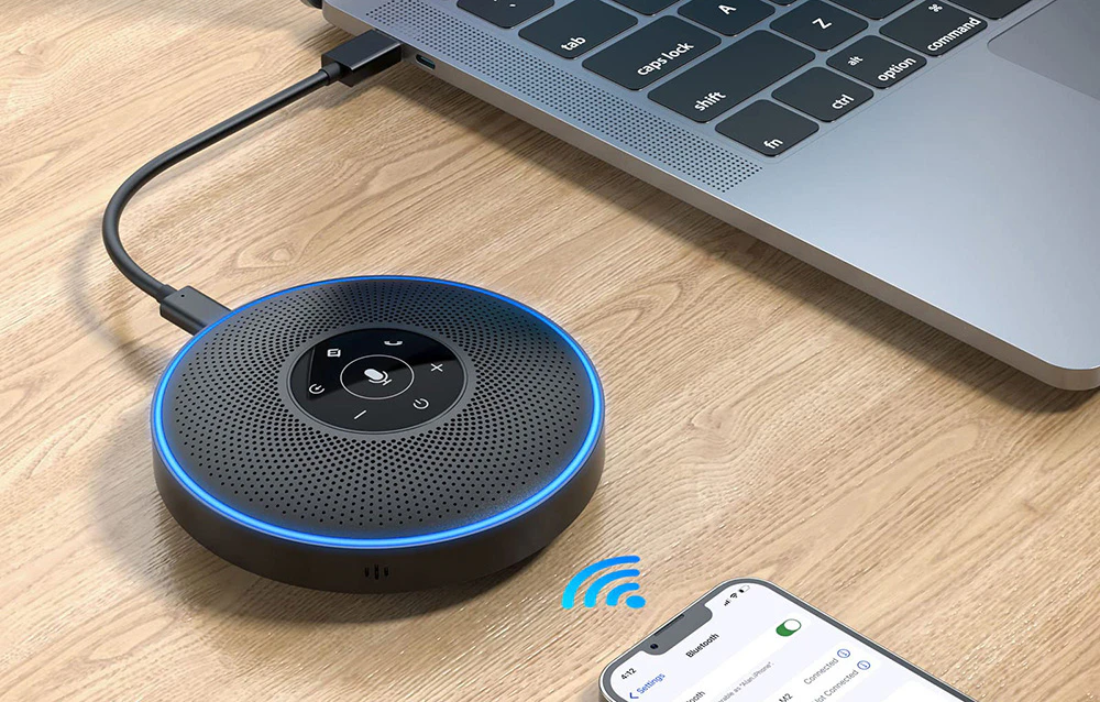 Un difuzor de conferință cu un inel luminos albastru conectat prin cablu la un laptop, comunicând simultan fără fir cu un smartphone prin Bluetooth