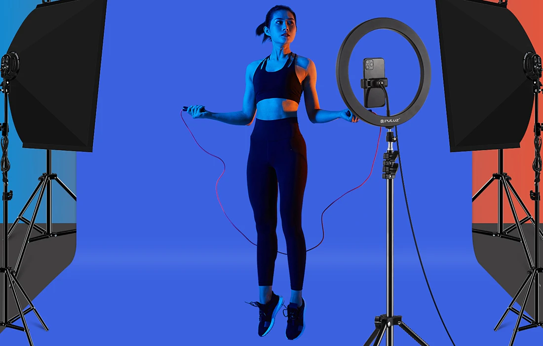O femeie în ținută sportivă sare coarda în fața unui fundal foto albastru, înconjurată de lumini softbox și de o lumină inelară cu un telefon