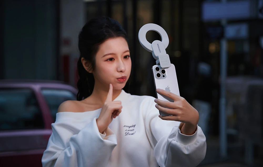 O femeie care își face un selfie cu smartphone-ul, având atașată o lampă inelară albă, pe fundalul unei străzi din oraș după apusul soarelui