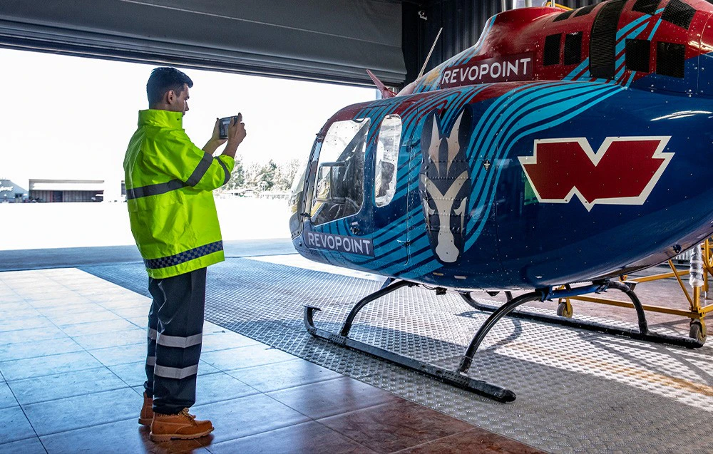 Un bărbat în jachetă de lucru verde scanează un elicopter.