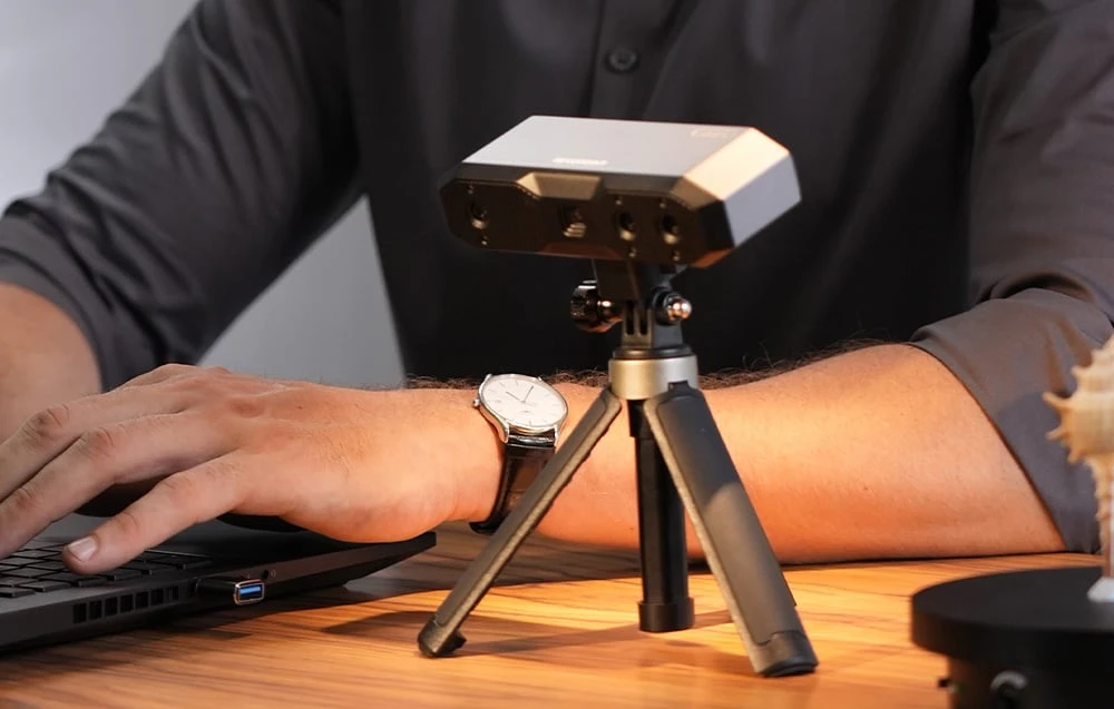 Scanerul 3D Revopoint Mini 2 pe un trepied lângă un bărbat care lucrează la un laptop pe un birou din lemn