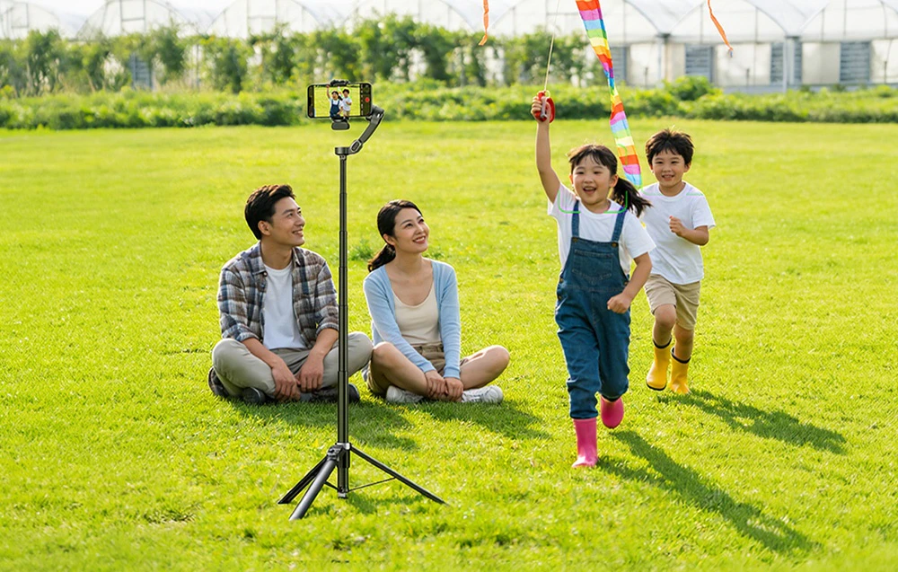O familie asiatică într-o grădină lângă un trepied cu un telefon care înregistrează, copiii alergând cu un zmeu