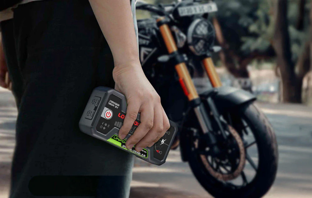 O mână ținând dispozitivul compact de pornire LOKITHOR pe fundalul unei motociclete parcate.