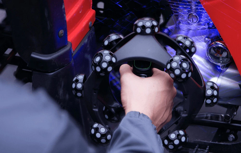 O mână care ține un scaner optic 3D portabil, scanând motorul unui vehicul roșu cu lumină albastră.