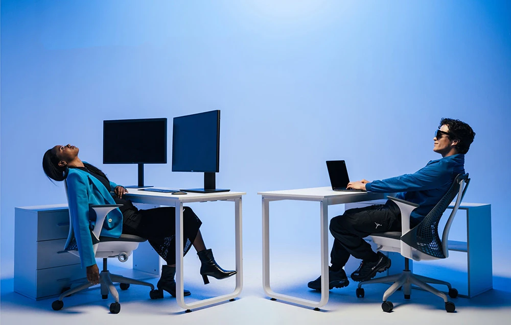 Femeie sprijinită pe un scaun la un birou cu două monitoare lângă un bărbat care lucrează la un laptop, ambele în lumina albastră a studioului