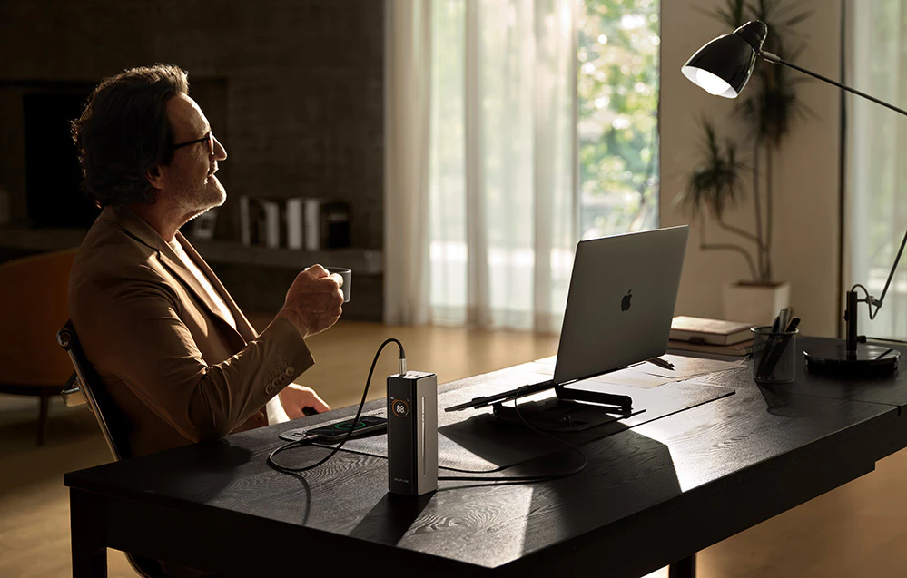 Un bărbat cu ochelari bea cafea la un birou cu un laptop și o lampă de birou într-o cameră luminoasă.