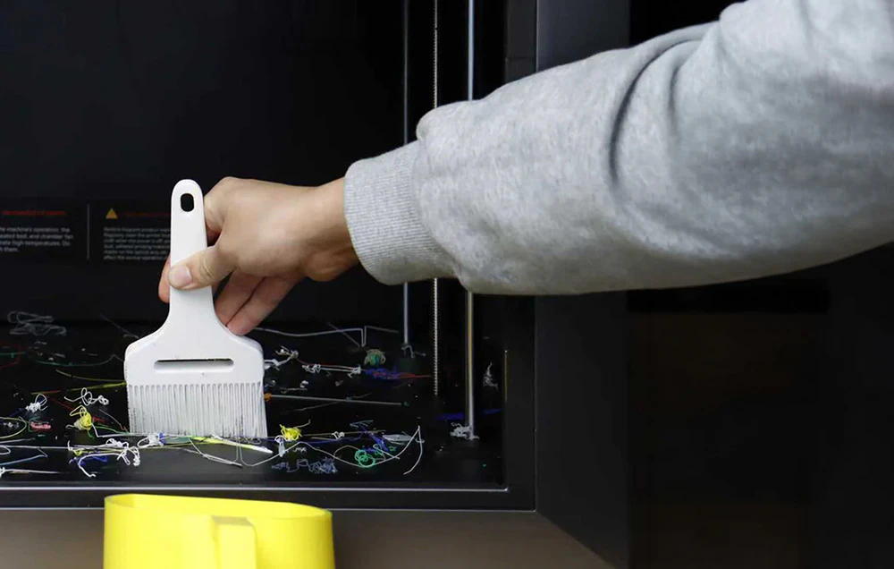 O mână îndepărtează reziduurile de filament de pe platforma de lucru a imprimantei 3D cu o perie.