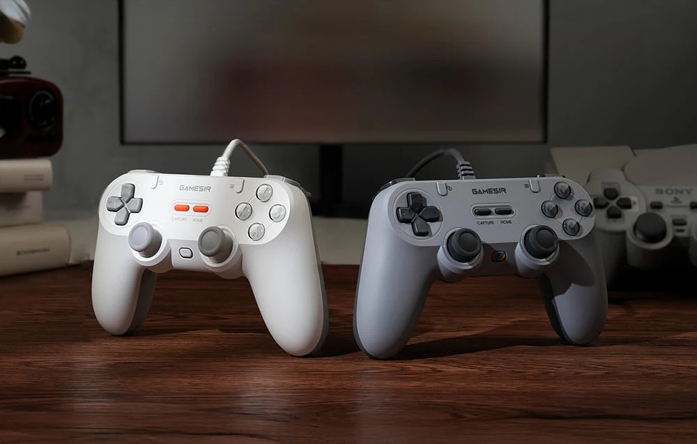 Două controlere GameSir cablate, alb și gri, așezate unul lângă altul pe o masă de lemn.