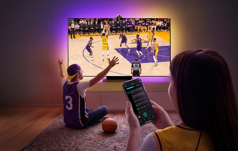 O femeie ține un smartphone în mână; un bărbat stă pe covor și urmărește un meci de baschet; peretele din spatele televizorului este iluminat în violet și galben