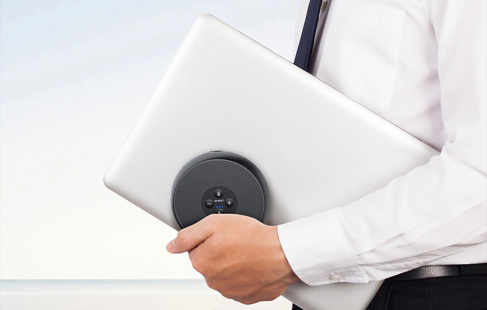 O persoană în cămașă albă și cravată ține sub braț un laptop argintiu închis, împreună cu un difuzor de conferință mic și rotund