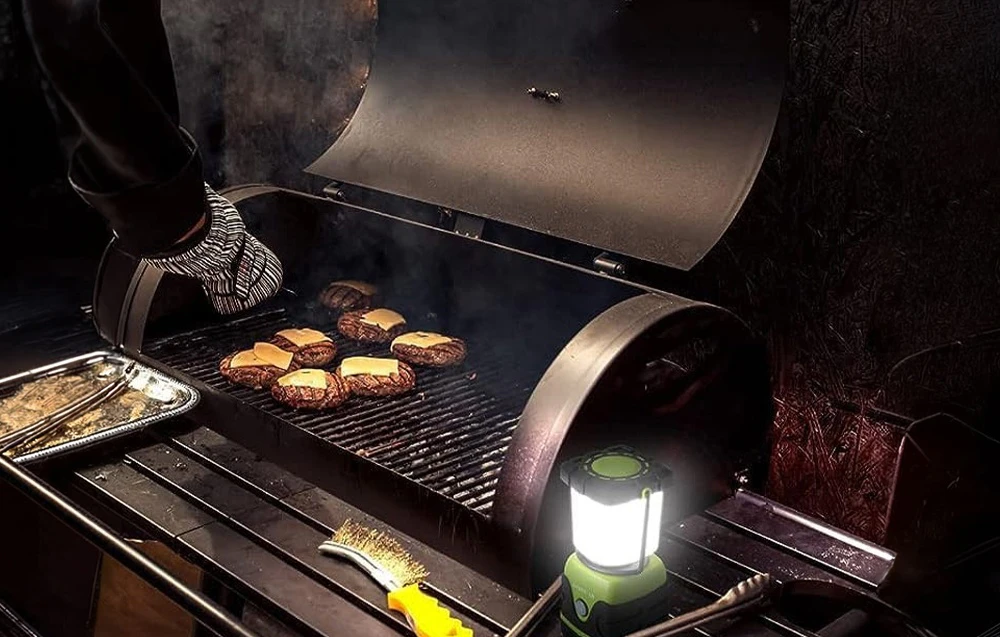 O mână într-o mănușă de grătar întoarce burgerii; o lanternă LED se află lângă grătar, iluminând ustensilele.