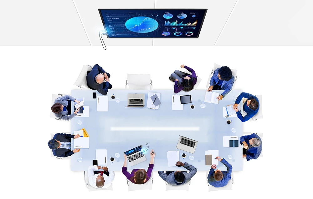 Vedere de sus a unui grup de persoane așezate la o masă mare de conferințe, cu laptopuri și un monitor pe perete