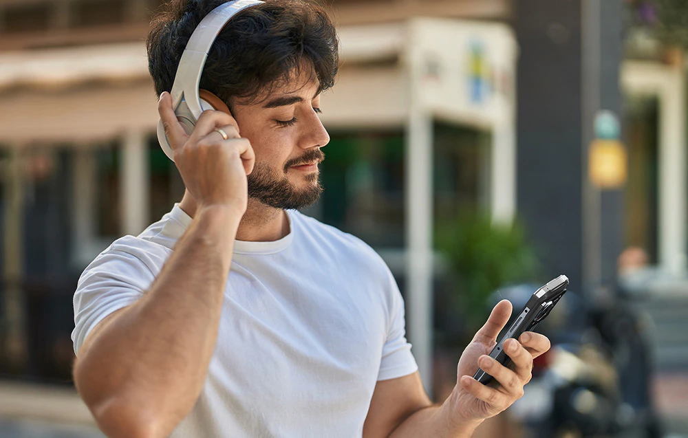 Un bărbat care poartă căști albe și utilizează smartphone-ul HOTWAV într-un cadru urban.