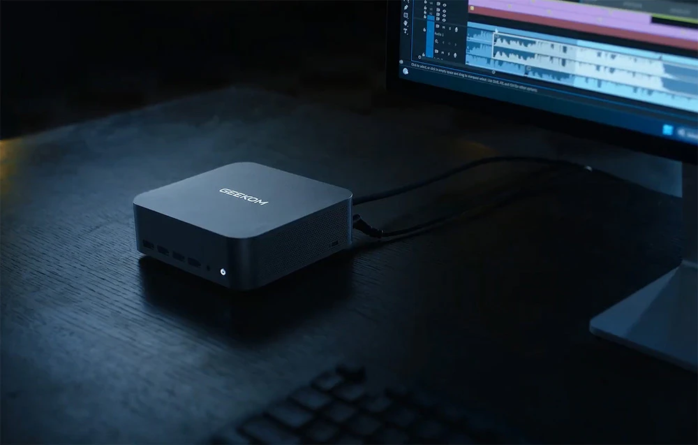 Geekom MINI PC pe un birou lângă un monitor într-o cameră întunecată