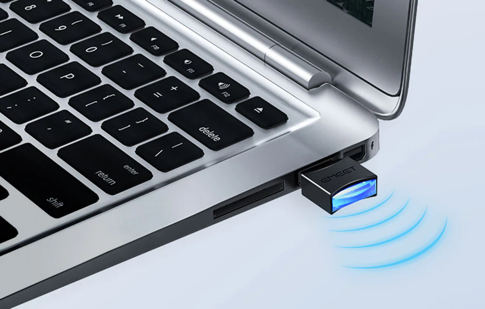 Prim-plan al unui receptor USB mic, negru, cu logo și iluminare de fundal albastră, conectat la portul lateral al unui laptop argintiu