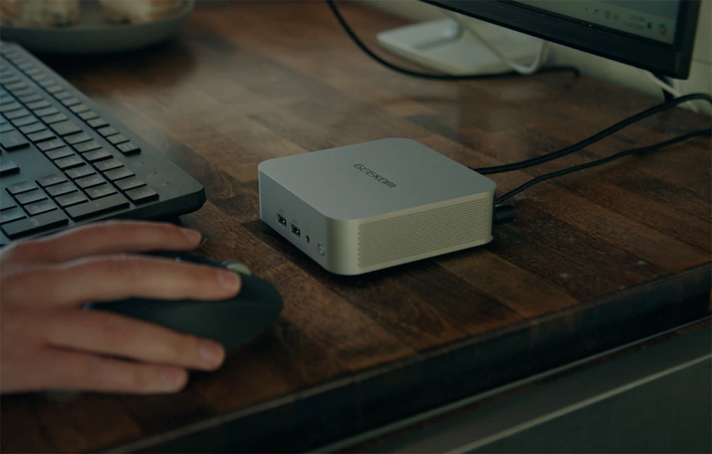 O mână ținând un mouse pe un birou din lemn, cu o tastatură și un mini computer alb lângă ea