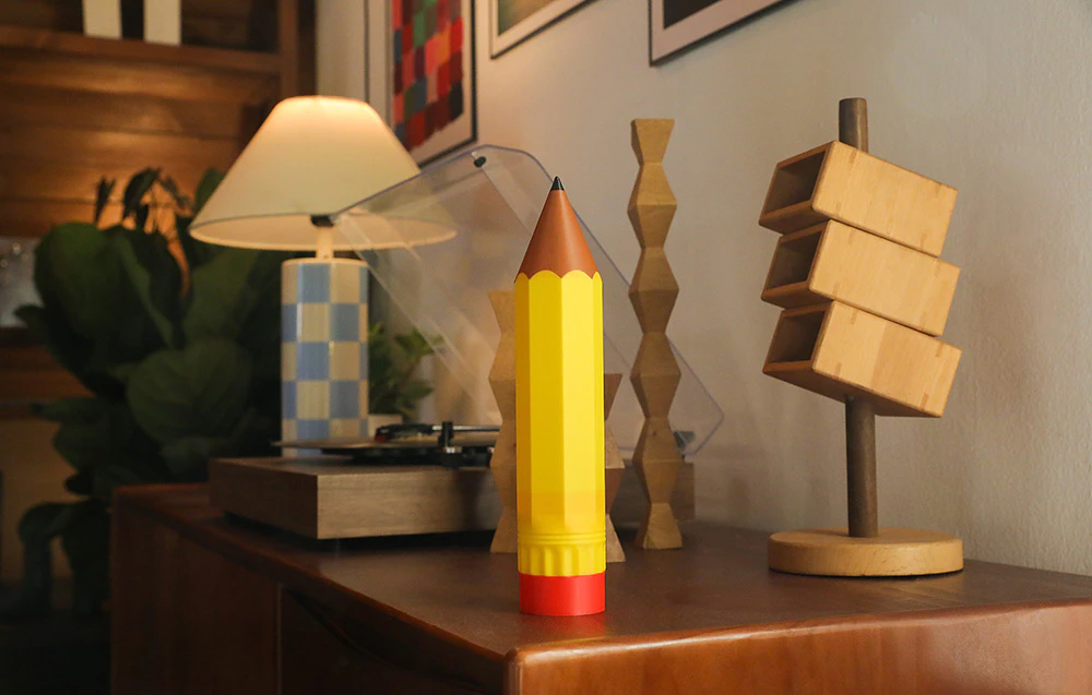 Un obiect mare în formă de creion galben stă pe o comodă lângă o lampă de designer și decorațiuni din lemn