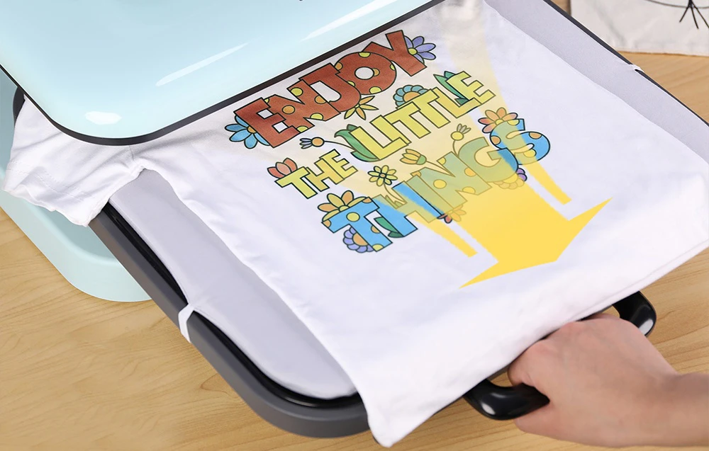Un tricou alb cu litere și grafice colorate fiind scos din partea inferioară a presei termice.